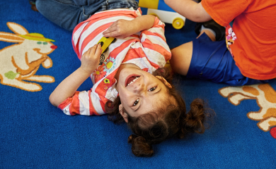 A child lays on her back on a rug while playing with a toy car