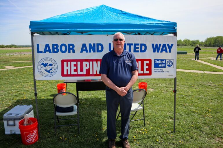 A person stands in front of a blue canopy tent with a sign reading "Labor and United Way, Helping People." The background features open grass and a clear sky. Nearby are chairs, orange buckets, and coolers.