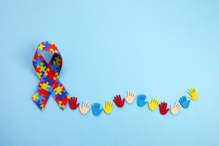 The image shows an Autism Awareness ribbon made up of multi-colored puzzle pieces. There are several paper cut-out hands trailing after the ribbon.