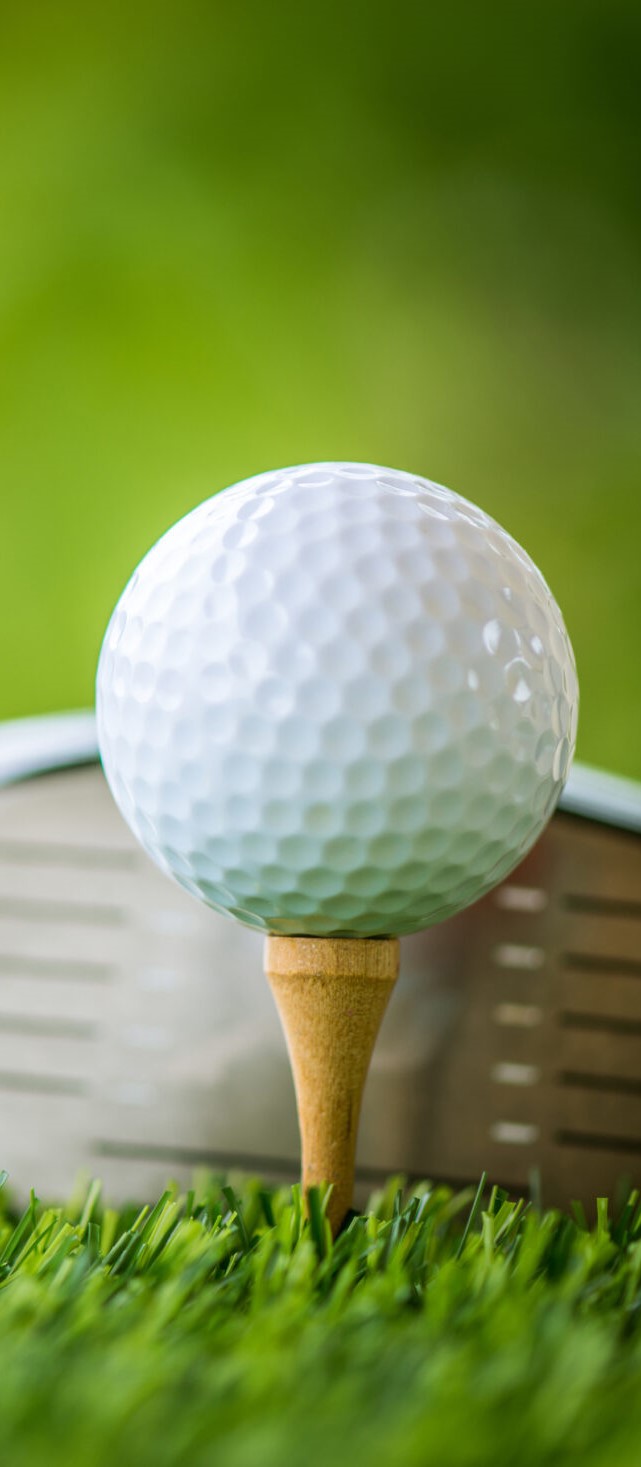 A golf ball on a wooden tee is set on a lush green grass field, with a golf club poised behind it, ready to strike. The background is blurred with shades of green and yellow, suggesting a warm day on the course.