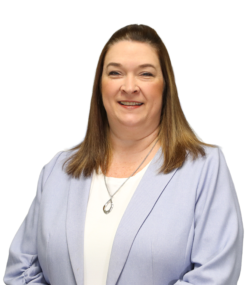 A woman with shoulder-length blonde hair smiles at the camera, exuding a sense of warmth. She is wearing a black jacket over a patterned blouse. The background features several blurred photographs on a wall, enhancing the friendly atmosphere.