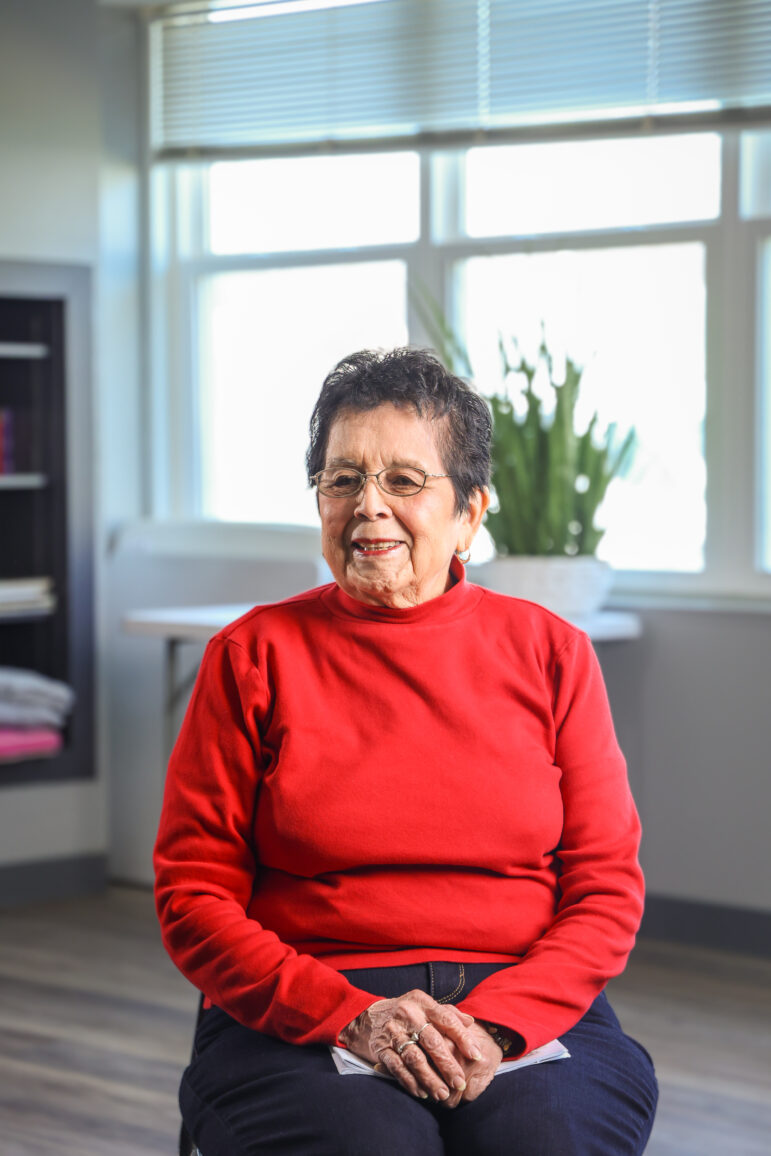 An older woman in a red sweater sits on a chair in the middle of a room, looking slightly away from the camera with a smile.