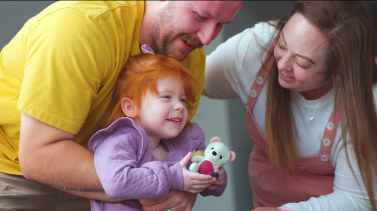 Two parents engage with their young red-haired daughter, who is holding a small stuffed teddy bear.