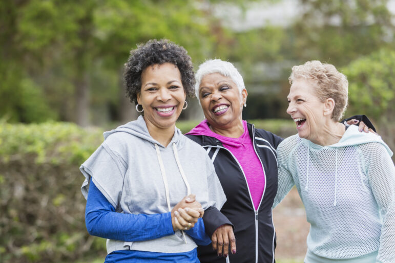 Three multi-ethnic women wearing hooded sweatshirts, hanging out together in the city, laughing while they take a walk. The woman with black hair, looking at the camera, is in her 50s and her senior friends are in their 60s.