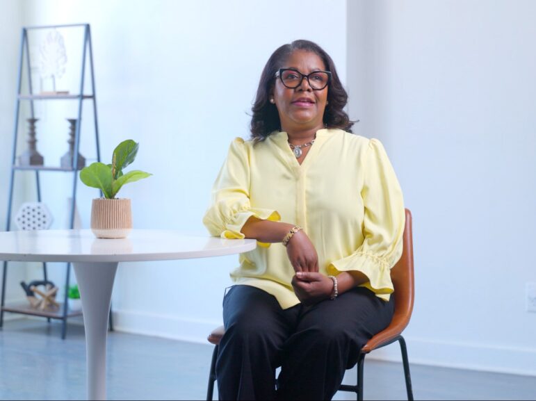 An African-American woman sits at a small white table in an orange chair. She is dressed professionally in a yellow button-down blouse and black slacks.