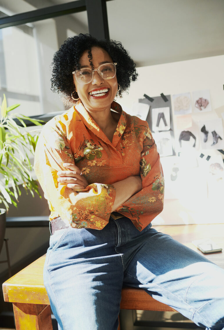 A mature black business woman wearing eyeglasses sits on her desk with her arms crossed while smiling.