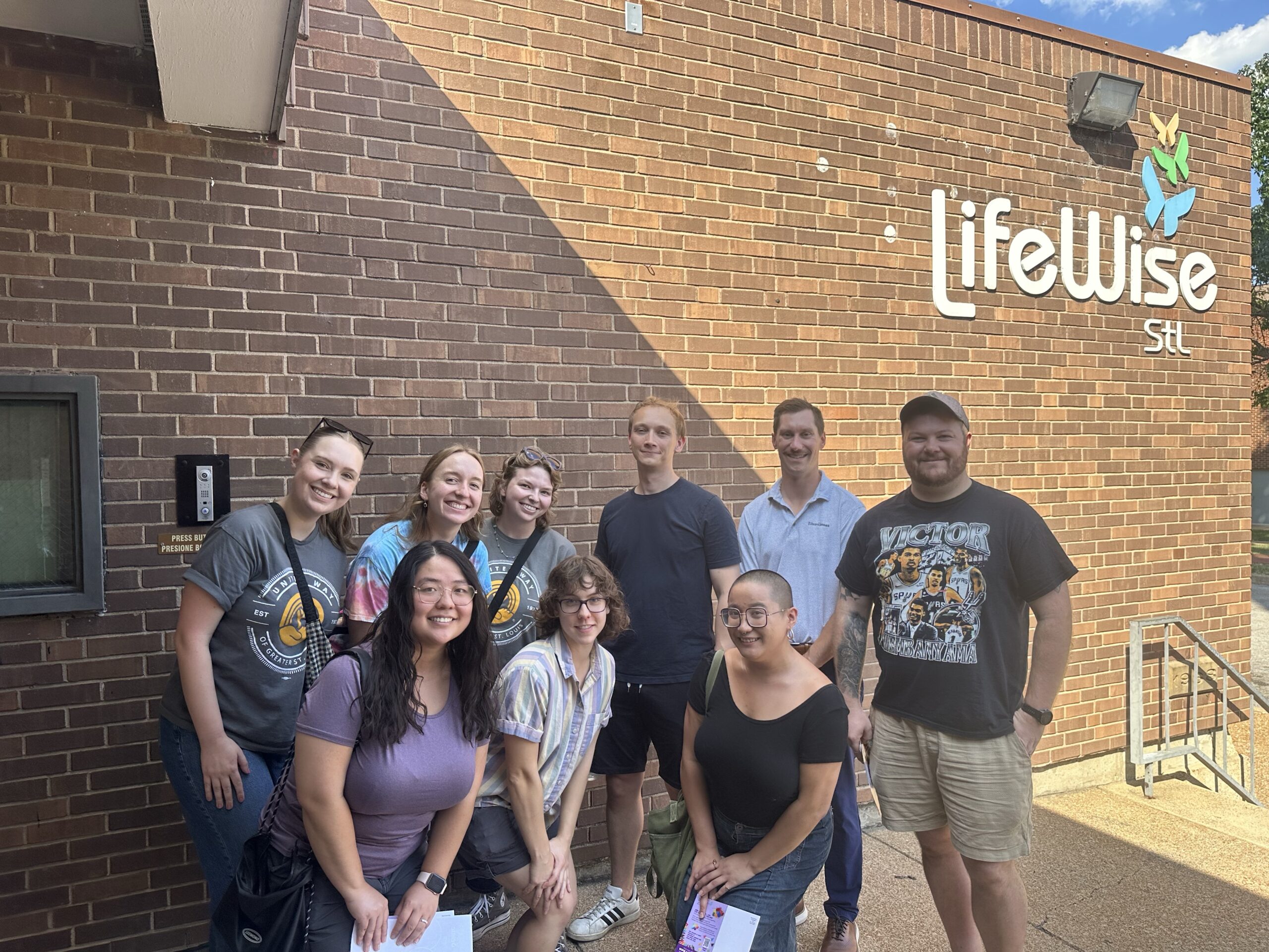 A group of people is smiling and posing in front of a brick building with a sign that says "LifeWise STL." They are wearing casual clothing, some sporting team-branded shirts. It is a cool, sunny day.